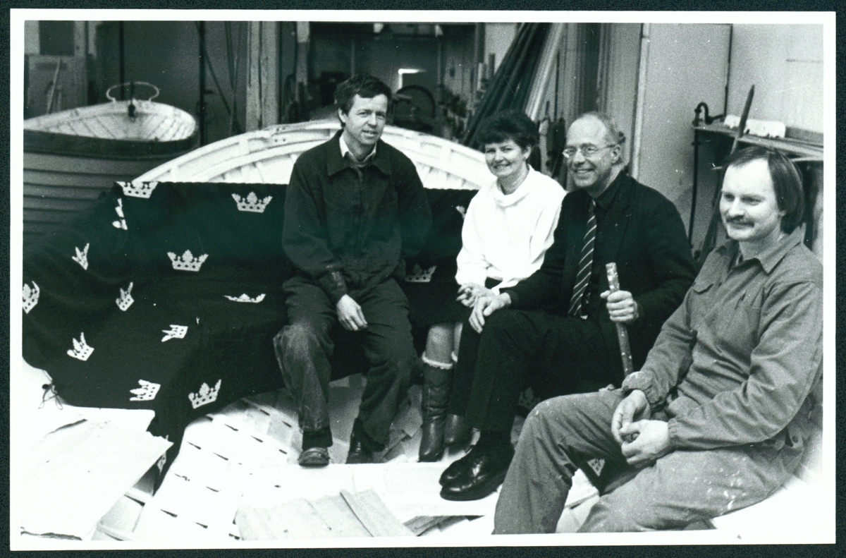 Bilden visar fyra personer sittande i en valbåt som har omkläds till kungaslupen nr 44 i Slup- och Barkasskjulet på Stumholmen i Karlskrona. De är från vänster: Krister Joelsson ÖrlB S, Karin Karlsson Sydosttvätt AB, Peter von Busch, MM, Kennet Svensson, Hästö Båtvarv. Fotot är tagen inför sjösättningen av kungaslupen 1988. Sittbänken i båten är belagt med tyg som är mönstrad med kronor.
