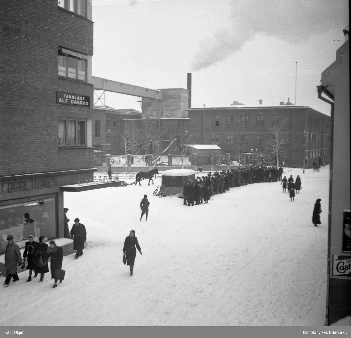 Folk i kø for kjøtt og fisk under 2. verdenskrig i Askim 1940-1945. Askim gummivarefabrikk i bakgrunnen.
Bygning til venstre, Bilitt, med skilt for tannlege Alf Dagrød, og glassmagasin i første etasje, ant. Askim Elektriske forretning og glassmagasin A/S.