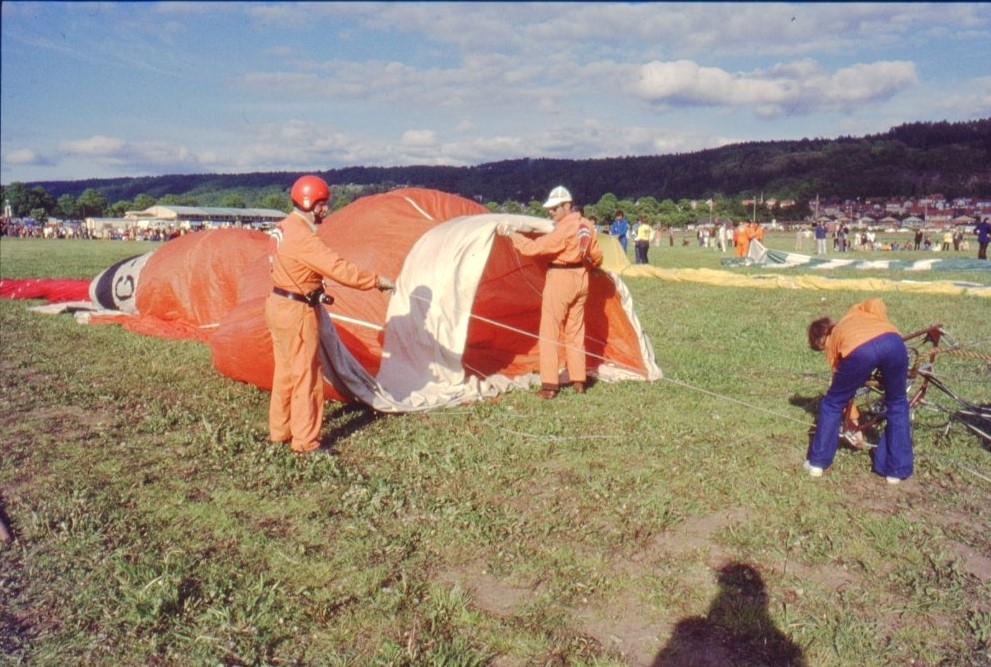 Två personer i orangea overaller med ballontryck på ryggen håller i öppningen av en ballong som håller på att fyllas med varmluft. En av personerna har en likafärgad hjälm på huvudet. En tredje person står hukad över brännaren.