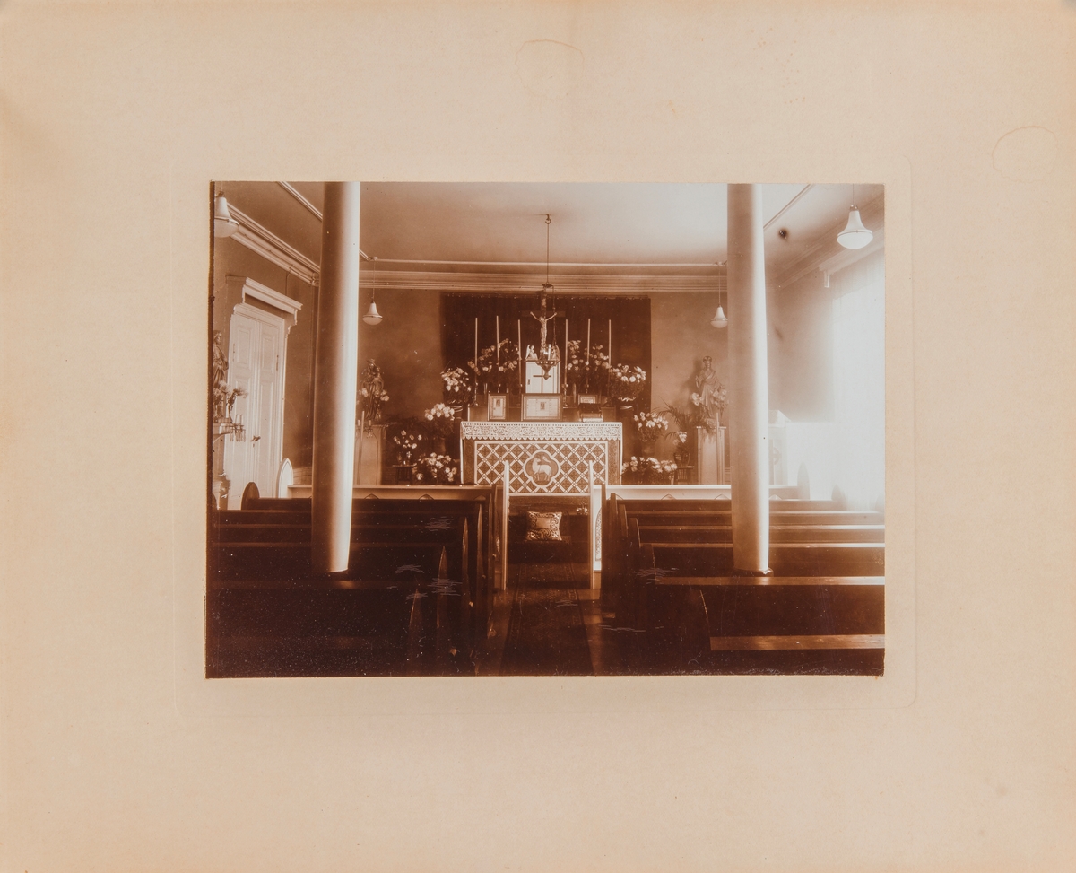 Bildet viser et kirkeinteriør med alter og et lite krusifiks omgitt av mye blomster. Det er ikke et typisk kirkerom, mer som en stor sal som er gjort om til kirke.
St. Torfinns kapell på Hamar.