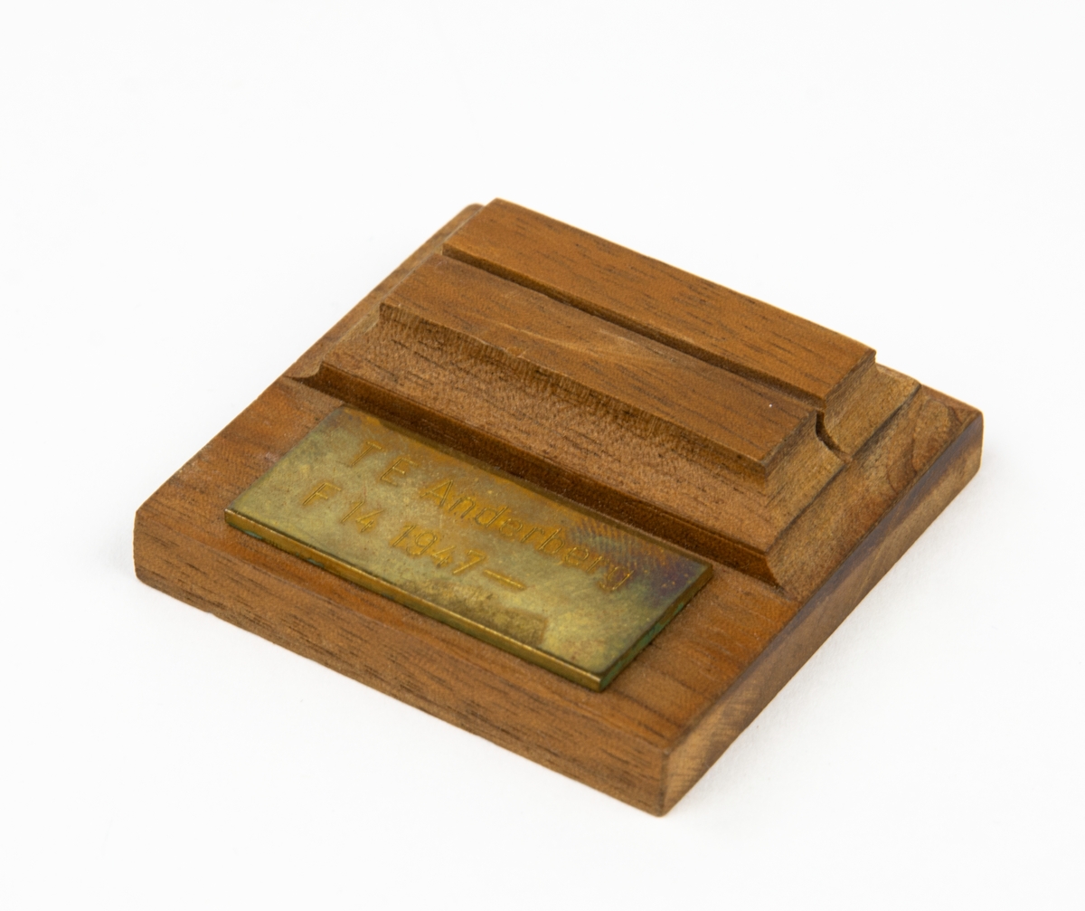 Kort/ eller papershållare med en skåra där kortet skall stå. På träbiten sitter en metallbricka med inskriptionen: T ANDERBERG F 14 1947-.