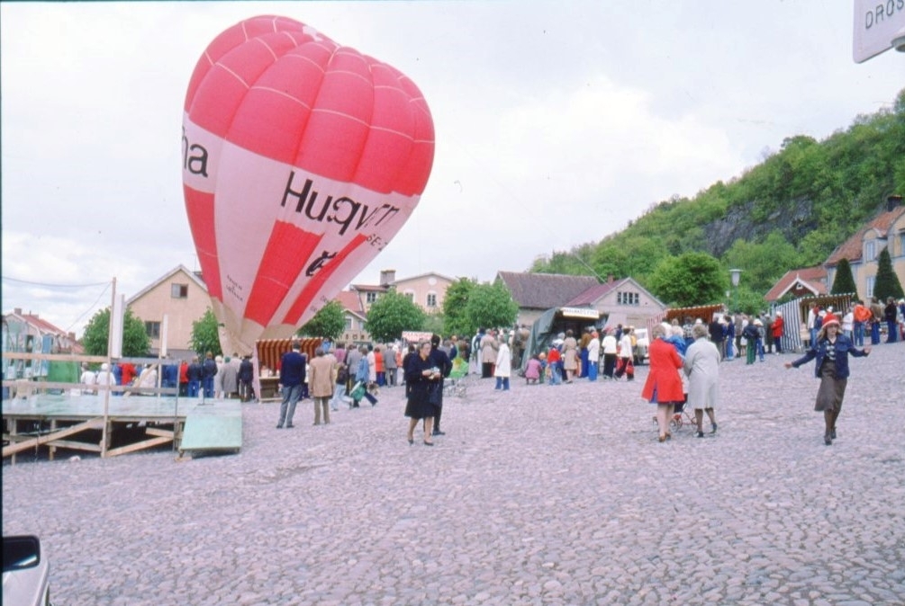 Den rödvita ballongen "Örnen II Gränna" med reklam för "Huskvarna" står fylld på Gränna torg intill en scen. Folk rör sig på torget.