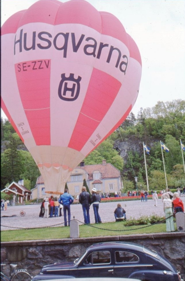 Den rödvita ballongen "Örnen II Gränna" med reklam för "Huskvarna" står fylld på Gränna torg. En liten folksamling runt ballongkorgen.