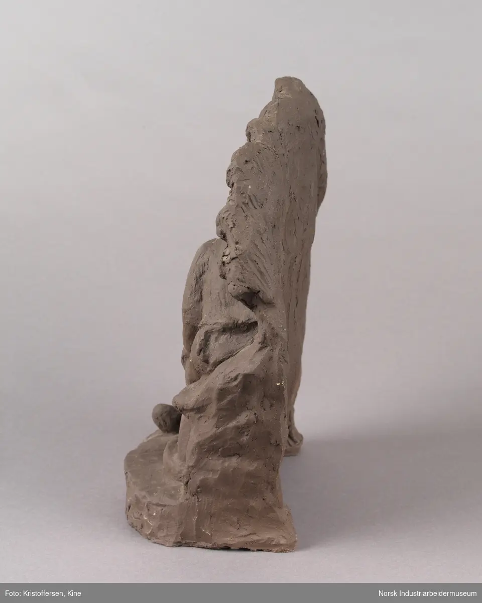 Skulptur laget av leire. En spelemann spiller på en fele, mens en tilhører sitter ved siden av. Tilhøreren har en hand på låret og luter hodet mot den andre handen. Skulpturen består av to sittende figurer, en grunn og en bakgrunn med bølgende kanter.