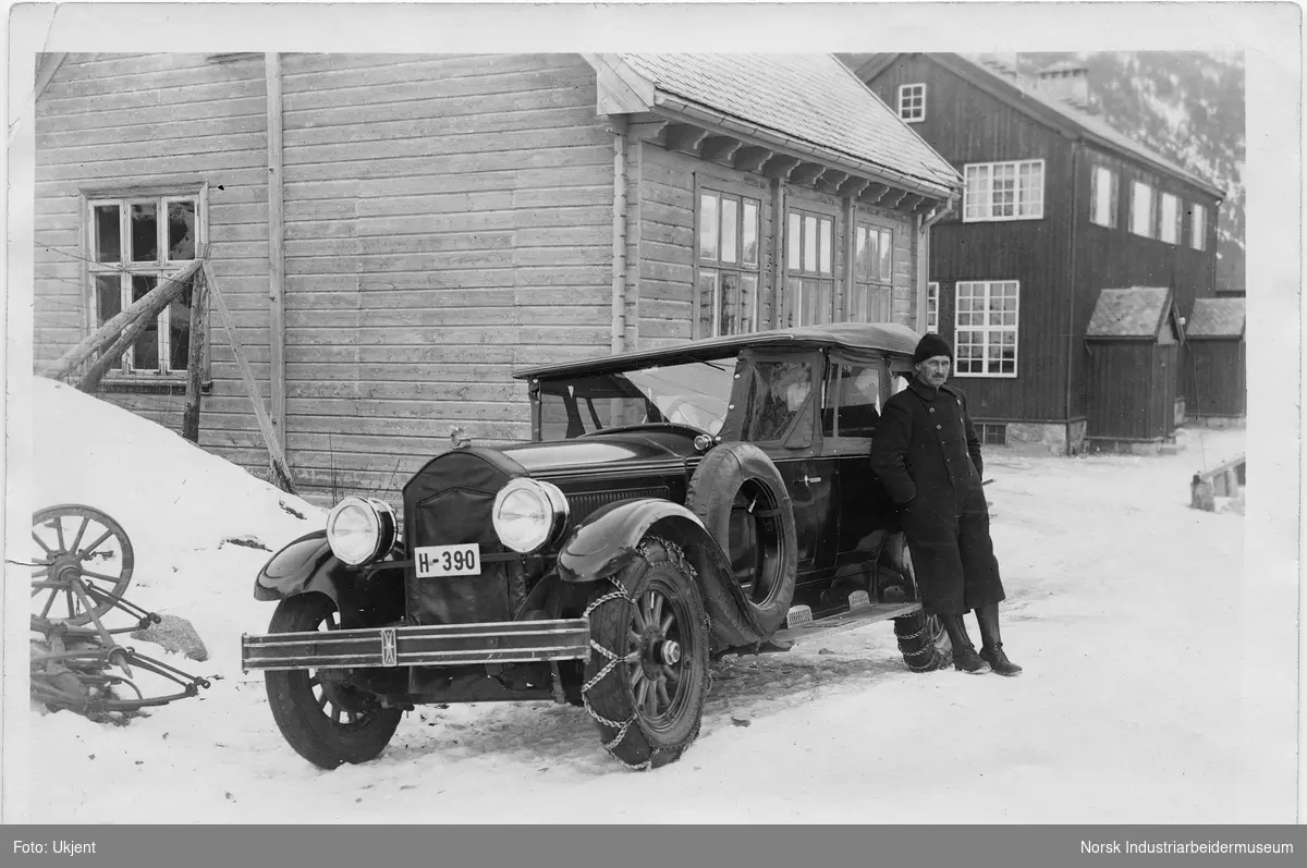 Mann i uniform stående ved siden av bil av merket Buick 1927 modell. Bilen har kjetting på et forhjul. Bilen eies av Rjukan Byanlegg, mannen er ansatt som sjåfør på Norsk Hydro. To hus i bakgrunnen.