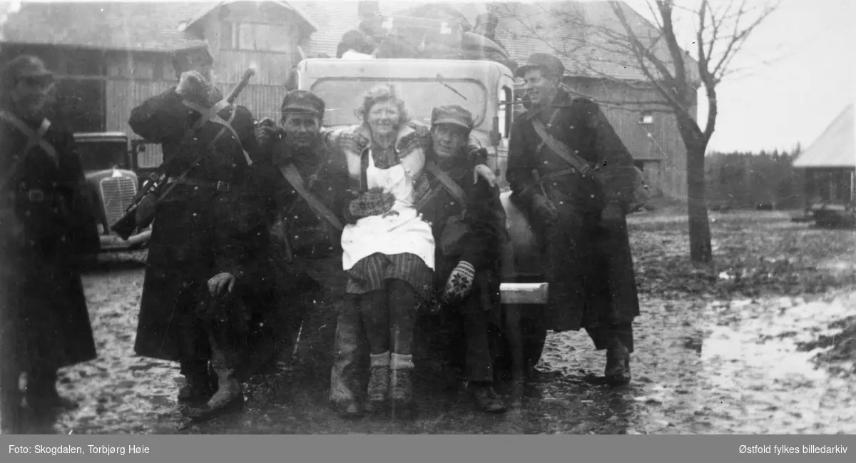 Fra gården Salmonrud i Eidsberg i april 1940, kaptein Rolf Graffs batteri under tilbaketrekning fra Fossum. Soldater og kvinne med kjole og kjøkkenforkle står foran person bil på tunet. I bakgrunnen uthus.