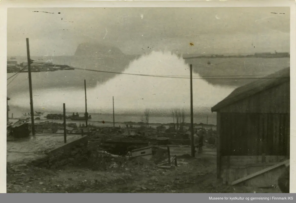 Detonasjon av en sjømine i havnebassenget i Hammerfest. Eksplosjonen finner sted ganske i nærheten av Molla-fjæra. I forgrunn ser man en del av en brakke og strømledniger. I midten av bildet ser man deler av hurtigrutekaia og fuglenes. I bakgrunn ser man Håja-fjellet i Sørøysundet.