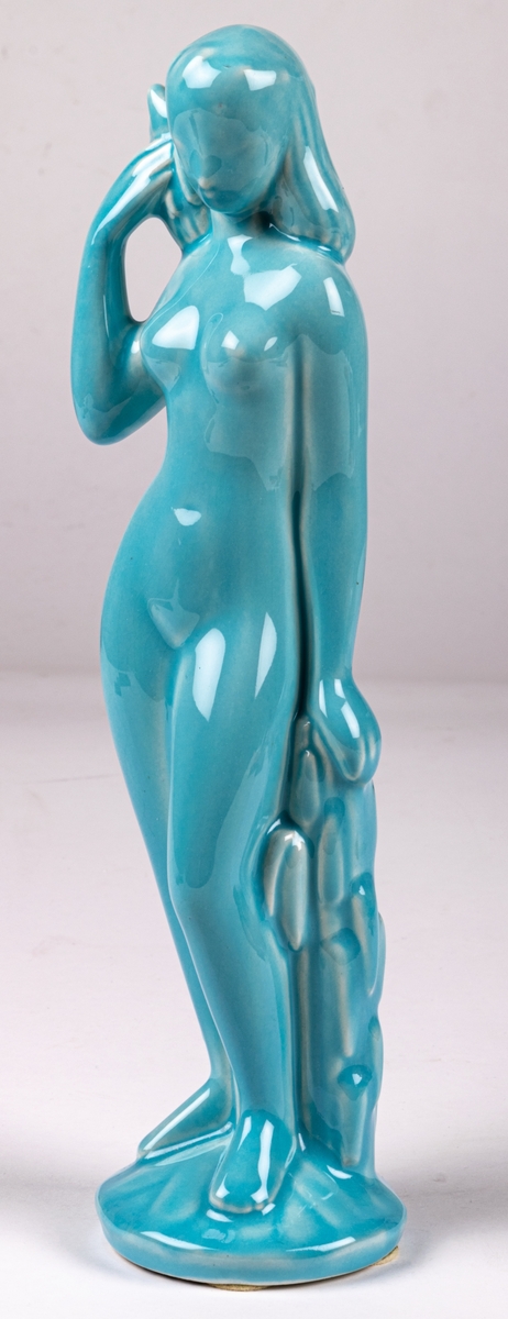 Figurin Eva, stående flicka, höger hand håller en korg med blommor på axeln, vänster hand lutad mot stubbe/buske. Posen och attributen ofta vanlig för att symbolisera ymnighet. Turkos glasyr. Formgiven av Eva Staehr Nielsen.
