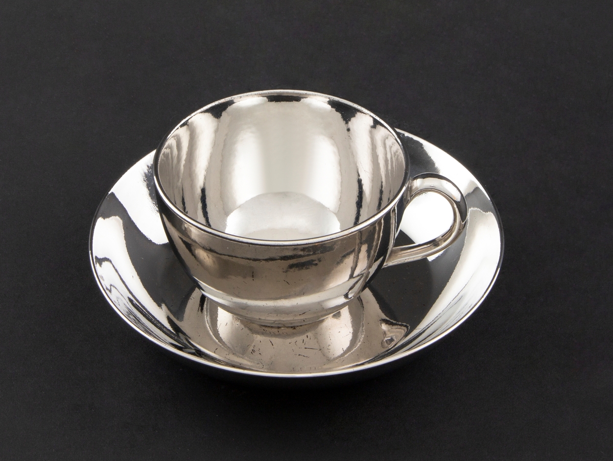 Kopp med skål i keramikk, glasert med sølvfarget luster. Tilnærmet sylinderformet korpus med påsatt hank. Lav sirkulær skål.