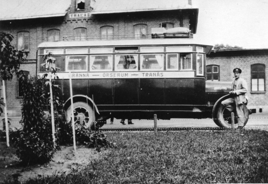 En buss står parkerad utanför stationshuset i Tranås. Framför bussen som är märkt på sidan: Gränna-Örserum-Tranås, står dess chaufför.