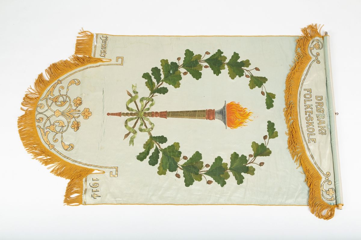 Skolefane. Silke. Hvitt stoff. 
Håndmalt dekor: Grønn eikeløvkrans rundt fakkel. Drøbak Folkeskole. 1914.
Stilisert rankemønster i bue nederst. Gyldengule silkefrynser. 
Fanen tredd inn på tverrstang av tre.