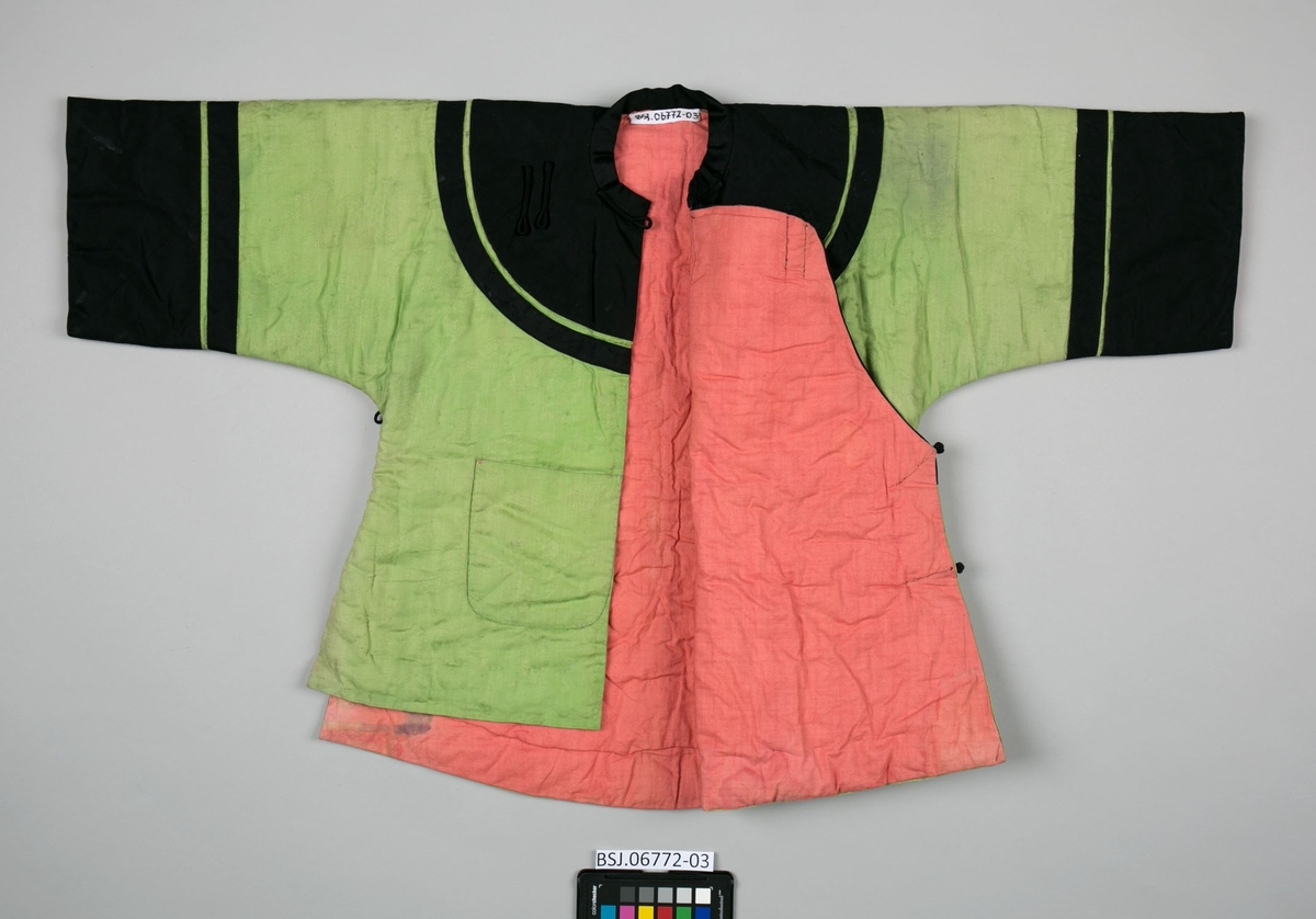 Kinesisk vattert omslagsjakke for barn (4-5 år) i grønn silkebrokade, kantet med svart brokadestoff. Lys rosa silkestoff i linningen (opprinnelig rød?). Lukkes med tre hemper. En utvendig lomme.