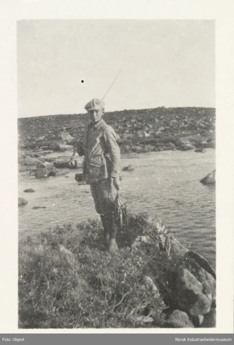 Gutt iført turklær og sikspens holder fiskestang og fiskefangst på Møsstrond. Bak sees innsjø og steinlandskap