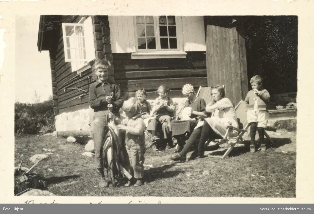 Kvinner i solstol og barn sittende på benk foran laftet hytte på Møsstrond. To gutter i front holder fiskefangst