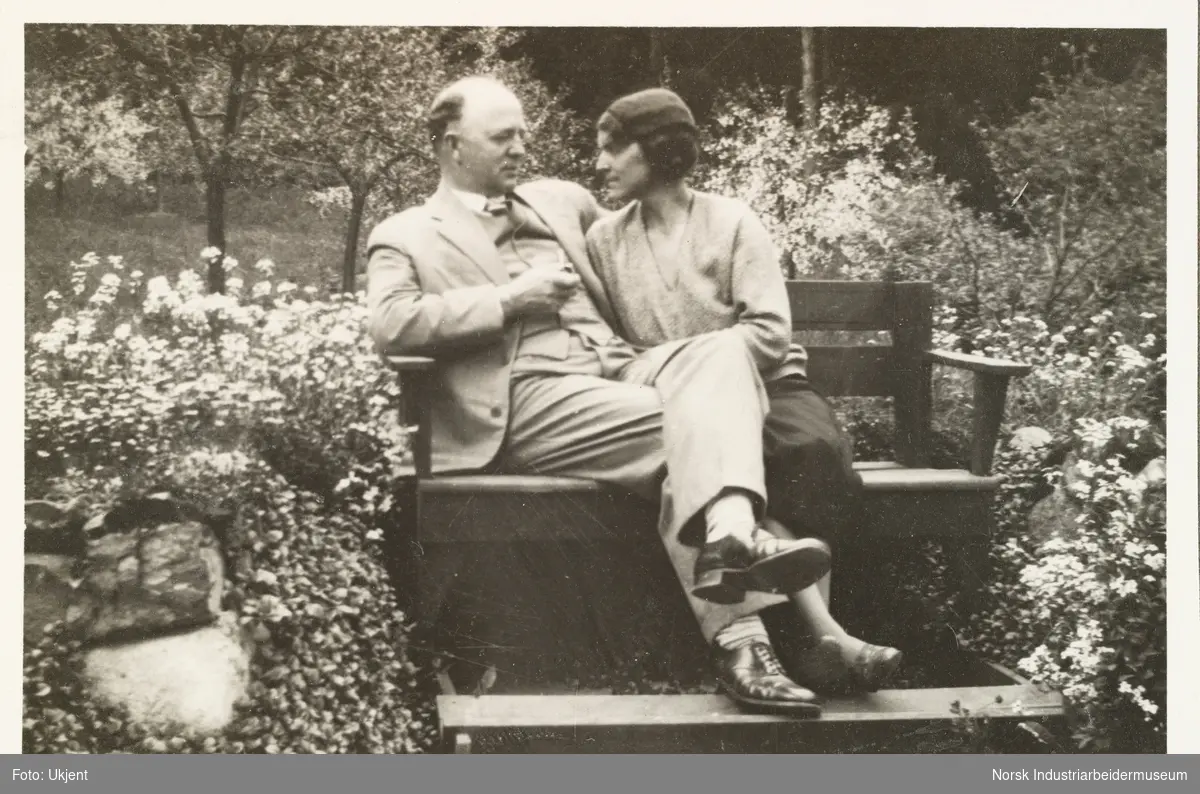 Høsten 1931. Kvinne og mann sitter på benk i hage. Kvinne lener seg mot mann. Mann har pipe i handen
