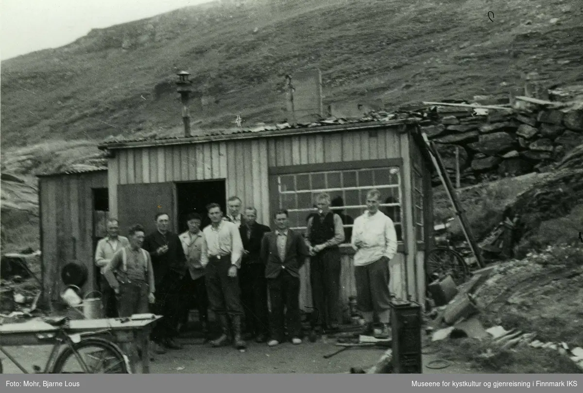 Gruppebilde av ti menn foran en brakke i Hammerfest. Mannen med hvit skjorte som står i midten, er Hans Heggelund. Til venstre står et bord og en sykkel. Til høyre av brakken ser man en mur og i bakgrunnen ligger Salenfjellet.
