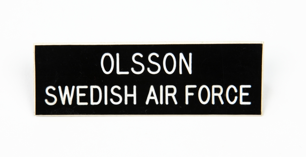 Namnskylt av plast med vit text på svart botten. På baksidan en fastsättningsanordning. 
Text: Olsson Swedish Air Force.