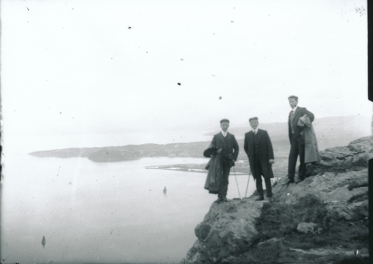 Tre unge menn iført frakker og samme hodeplagg, sannsynligvis studenterluer, står på toppen av høyt fjell. Hav og kystby i bakgrunnen.