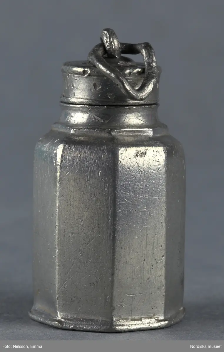Inventering Sesam 1996-1999:
H 5,5 (cm)
Flaska, till dockskåp, tenn, åttkantig, skruvlock med ögla. Saknar stämplar.
Tillhör dockskåp 151.825.
Bilaga
Anna Womack 1996