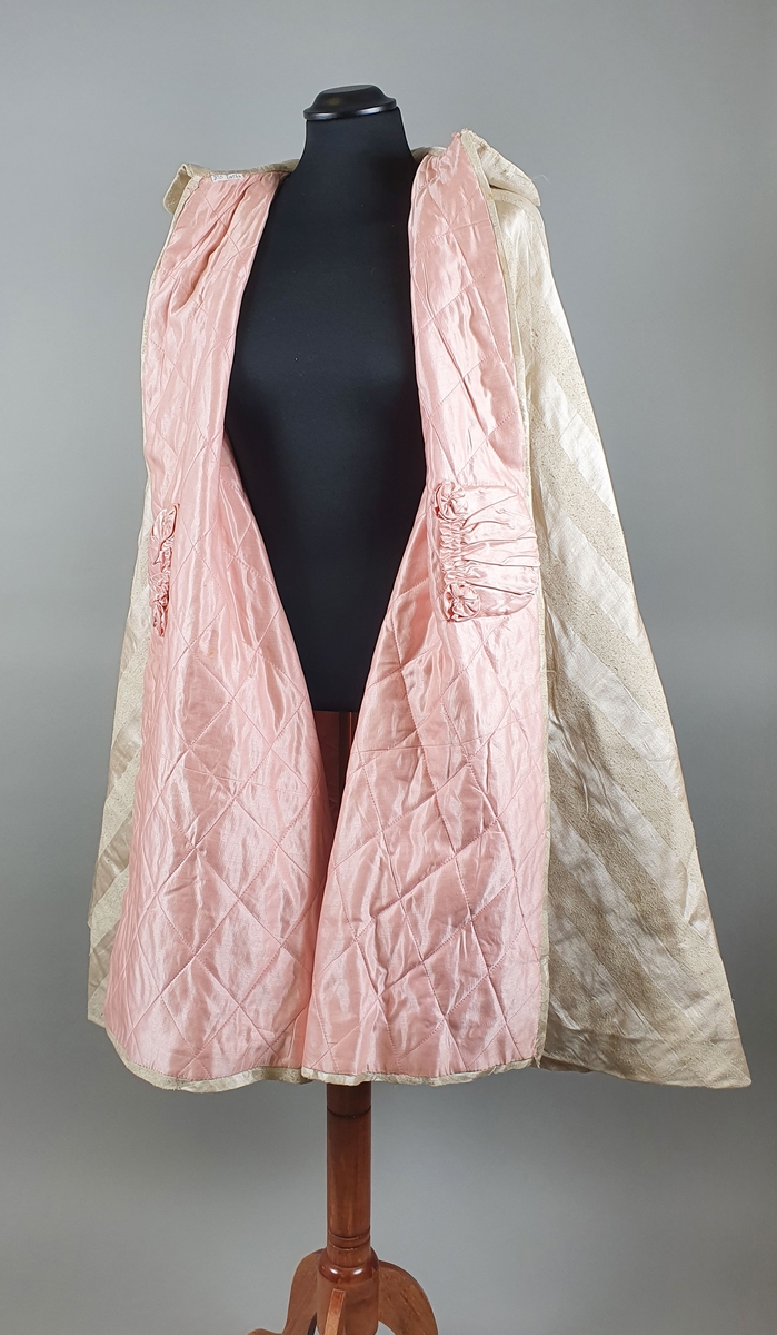 Hvit cape med diagonale striper vevd i lin og silke. Rosa fôr av bomull. Capen er vattert. To lommer på innsiden av hvert forstykke. Bred skjortekrage med buede kanter.