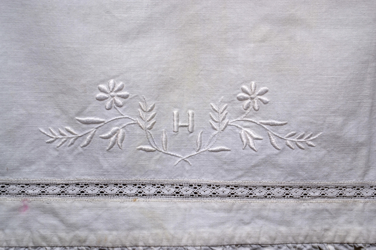 Pyntehåndkle i bomullstekstil med innfelling og avslutning i industriprodusert blondebord. Håndbrodert monogram det brodert et monogram H og blomster i plattsøm.