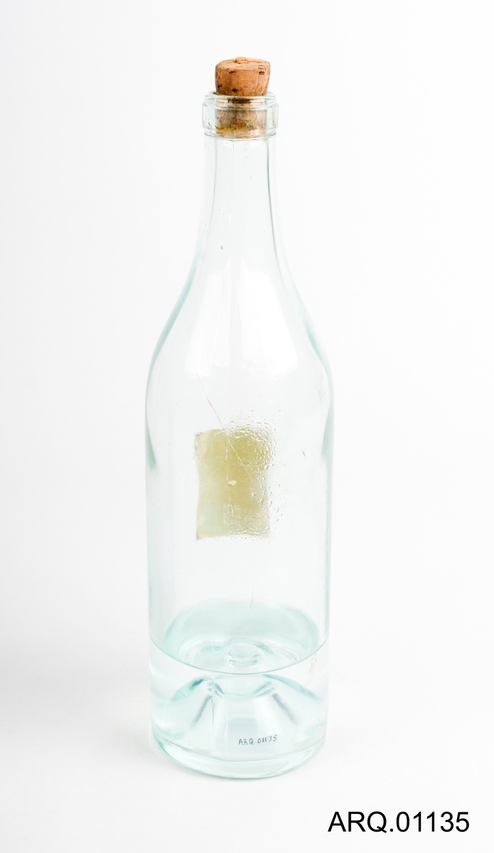 Gjennomsiktig glassflaske med blå-skjær. Liten Etikett med rød og svart skrift. Korken sitter i og det er blankt innhold i flasken.