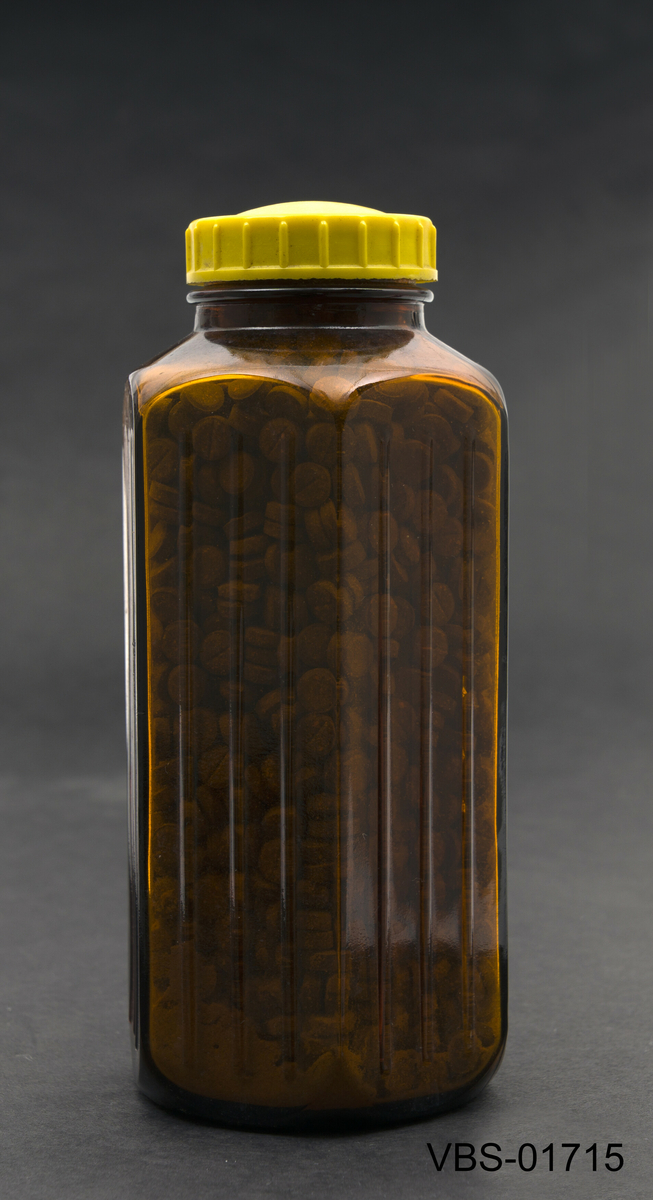Sekskantet laboratorieglassflaske i ravbrun farge. Nederst på flasken er det en lettelse med påskriften 500.
Inneholder brune tabletter.
Gul bakelitt skrulokk.