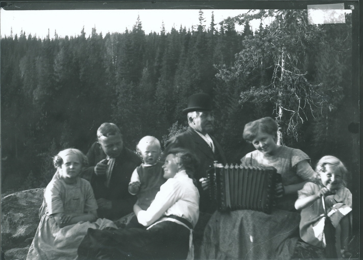 Medlemmer og venner av familien Rydgren sitter på en fjellrabb med skog i bakgrunnen. En av kvinnene i gruppen spiller trekkspill. Bestyrer Wilhelm Rydgren er iført skalk og dressantrekk.