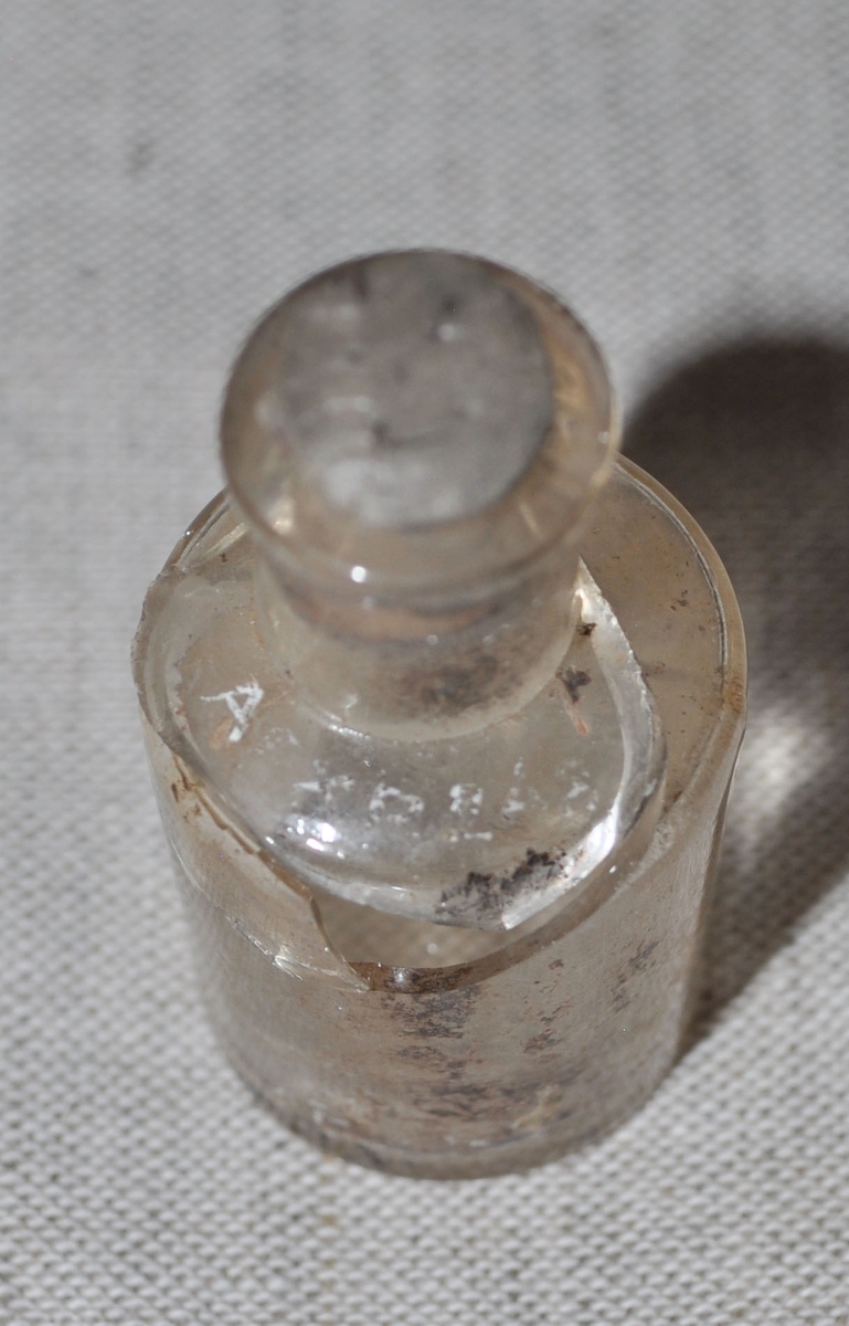 Ovandel av en genomskinlig glasflaska. Det siter en liten kork i flasköppningen. Trasig.
Den andra delen av flaskan har ett annat nummer (APXP.299)