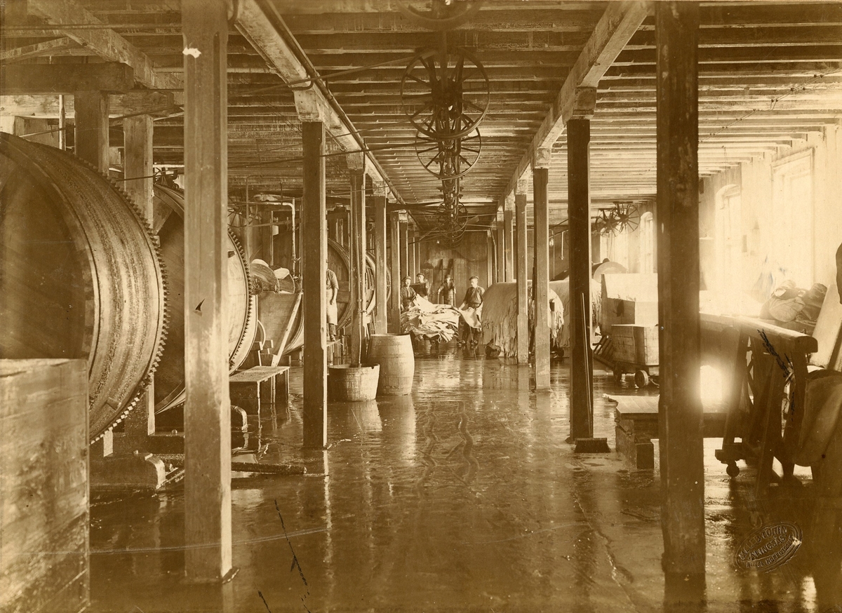 Bilden visar falsverkstaden på Göta läderfabrik, kv. Syrenen. Stora trähjul till vänster samt 4 arbetare, längre in, som står bakom en hög med skinn. Text på baksidan "Långelyckan Andra våningen".