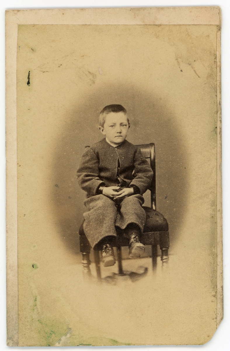 Portrett av en liten gutt, Eilert Brodtkorb (1862-1931). På bildet er han ca. 5 år gammel og sitter på en stol. Bak stolen kan vi se foten til nakkestøtten. Gutten er kledd i en mørk dress. Dette er et av de eldste bildene i fotoarkivet, tatt av Olaf Olsen ca. 1867. Fotograf Olaf Olsen (Trondheim) var aktiv i perioden 1864-1883.