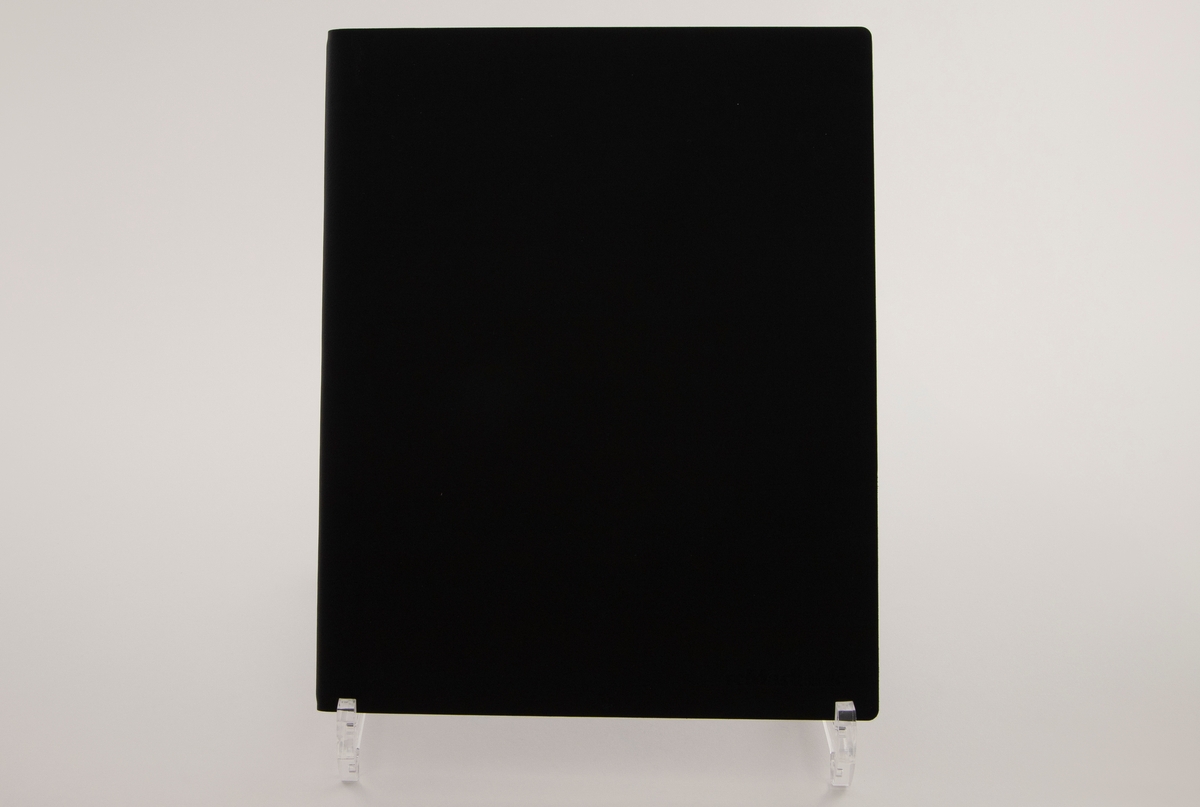 Rektangulært sortfarget deksel i lær, som oppbevares i papiremballasje med grålige og sorte fargenyanser.