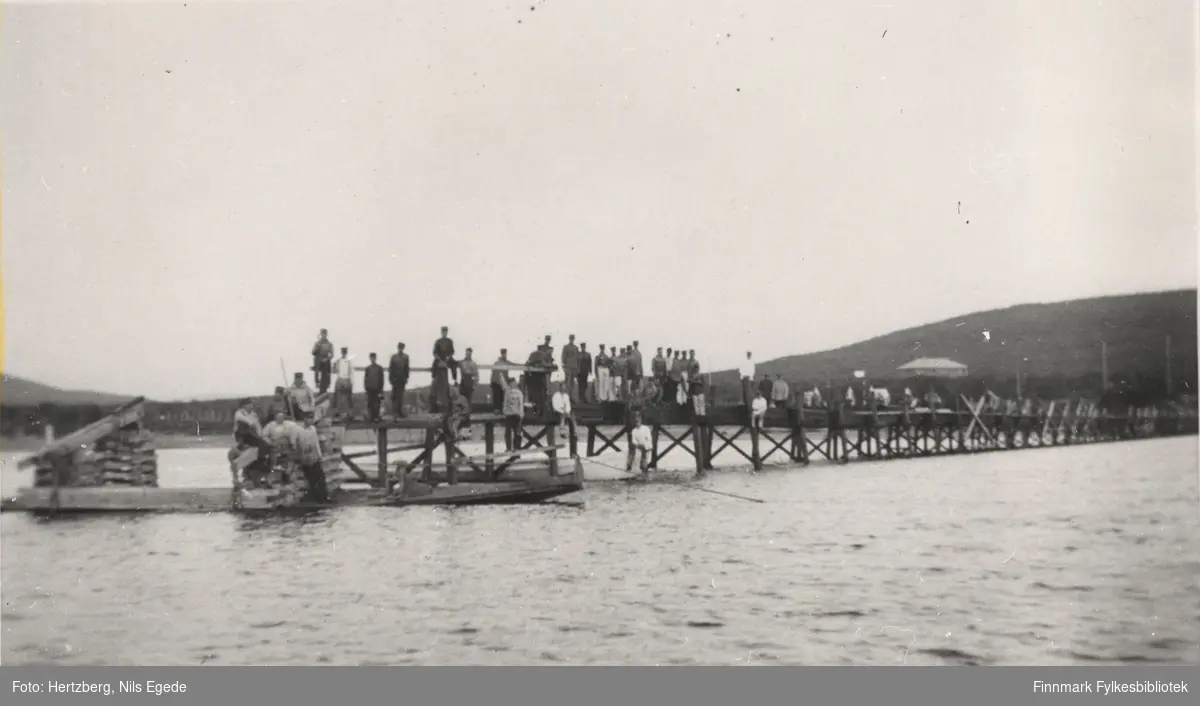 Brubygging over Tanaelven i Seida. Mange mennesker står eller sitter på den halvferdige bruen. Ennå en av bukkene er kommet på plass. Seida-tana, 1939.