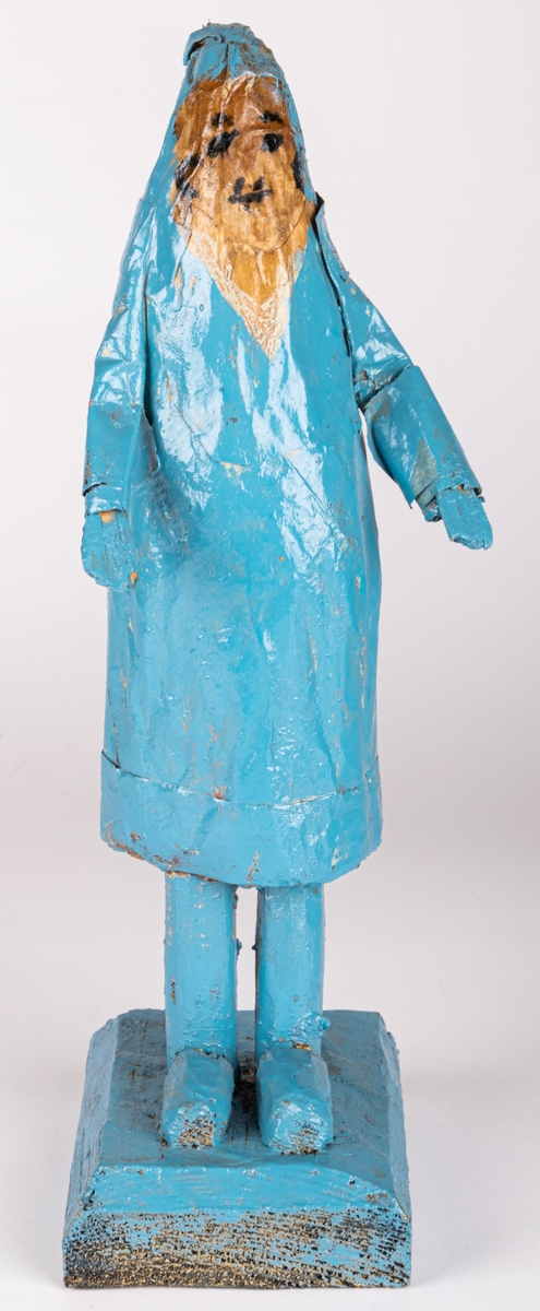 Skulptur av trä och papper. Figur/människofigur, helt blåmålad. Ansikte i beige med detaljer i svart. Mössliknande form på huvudet, med garntofs bak.