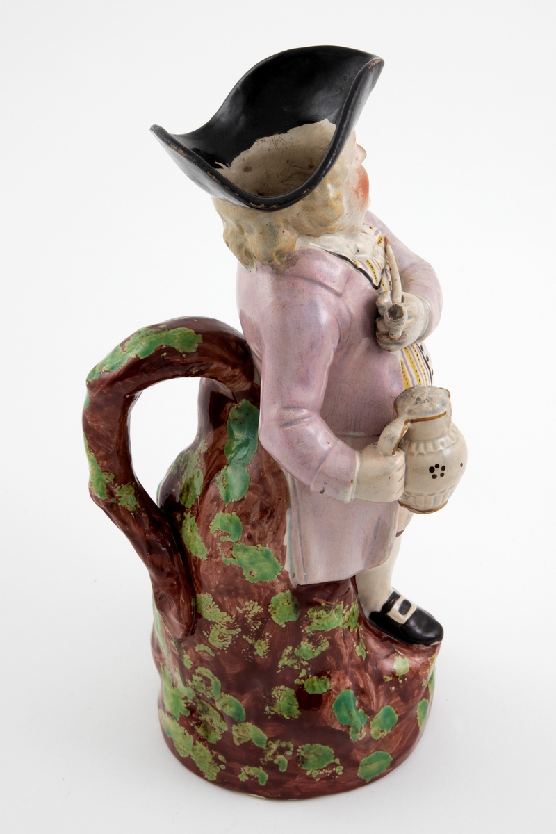 Toddykanne i form av en stående mannsfigur med pipe og kanne i hendene. Lokket mangler. Dekor i brunt, grønt, svart, gult og fiolett.