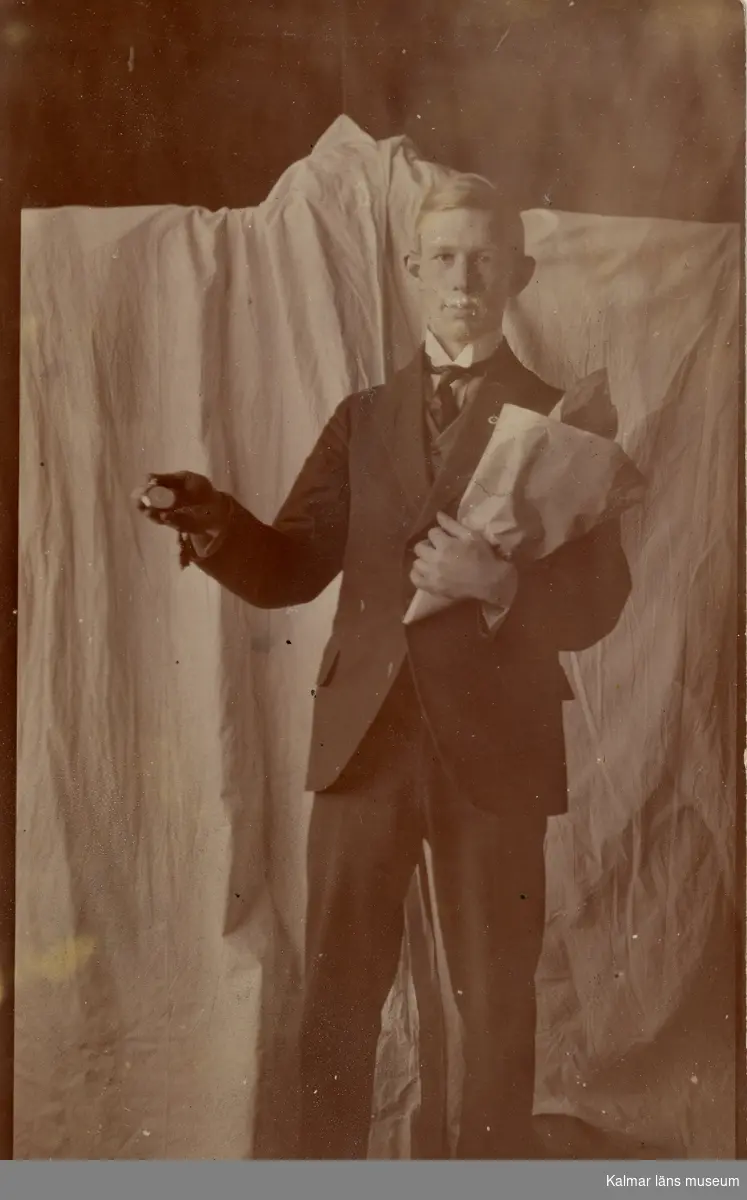 Fotografi föreställande Curt Nyström Stoopendaal. Han är avbildad framifrån och är klädd i vit skjorta, mörk slips, väst, kavaj och byxor. Han poserar framför ett vitt skynke. I ena armen bär han papper som kan innehålla en bukett med blommor i den andra handen håller han upp en klocka ett fickur. Han har mustasch.