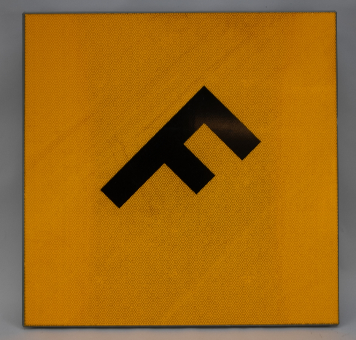 Rätvinklig rombformad tavla med bockade kanter. På baksidan finns fästen för stolpe. Framsidan är täckt av gul reflexfolie och har bokstaven "F" i svart i mitten.