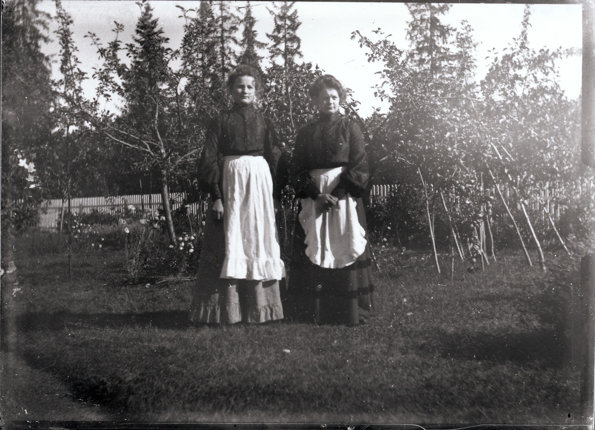 To kvinner i lange kjoler med forklær over står i hage med frukttrær, bærbusker og blomsterbed. Stakittgjerde i bakgrunnen.