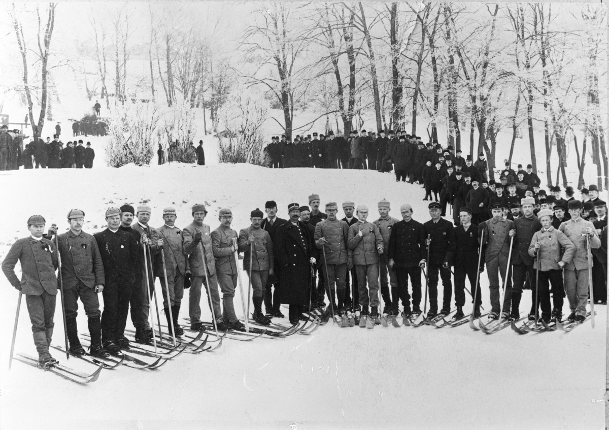 Menn i uniform og ski oppstilt i snødekt skog.