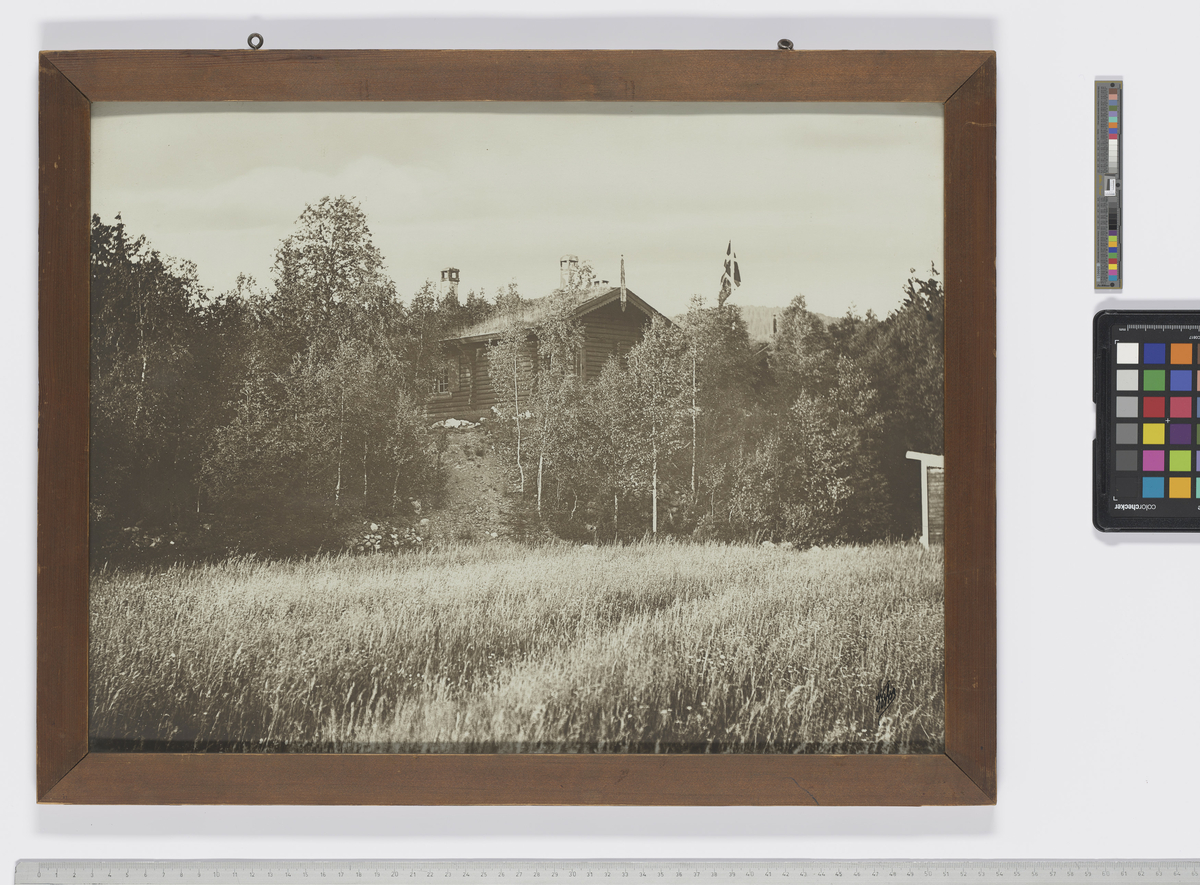 Landskapsbilde med skog, et jorde i forgrunnen. I midten av bildet sees en bygning, med det norske flagget heist opp.