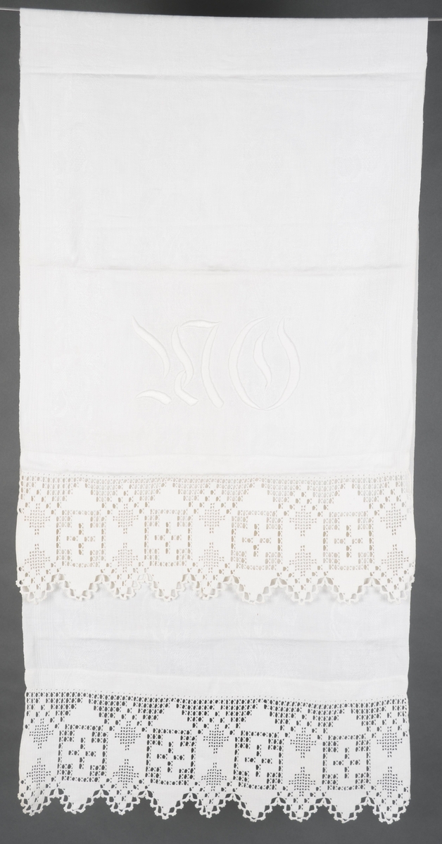 Hvit pyntehåndduk hvor kortsidene er brettet inn og sydd til med maskin. Heklet bord er sydd på for hånd i hver kortside. Broderte initialer i tykksøm.