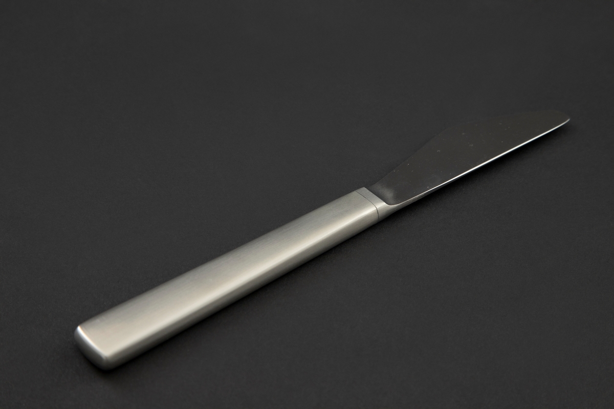 Bordkniv i rustfritt stål. Knivens blad har en avrundet trekantform med små tagger på øvre kant og rett linje fra bladrygg til skaft. Linje ved overgangen fra blad og skaft. Glatt overflate.