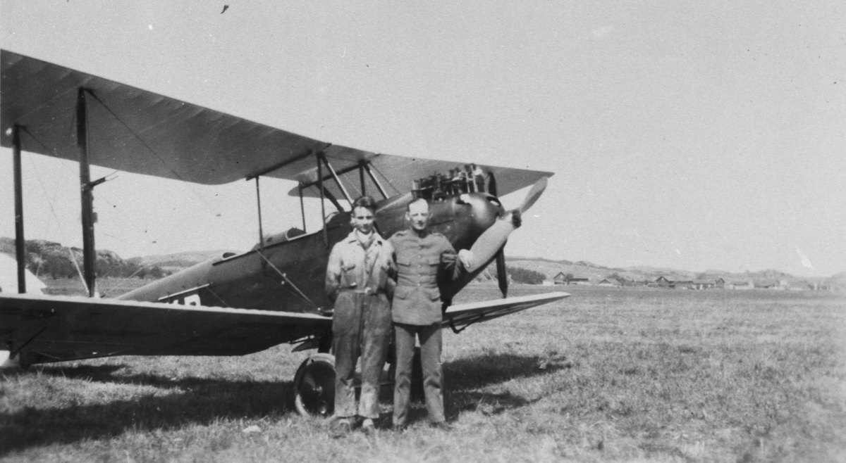 Två oidentifierade militärer står framför flygplan DH. 60 Moth på ett flygfält.