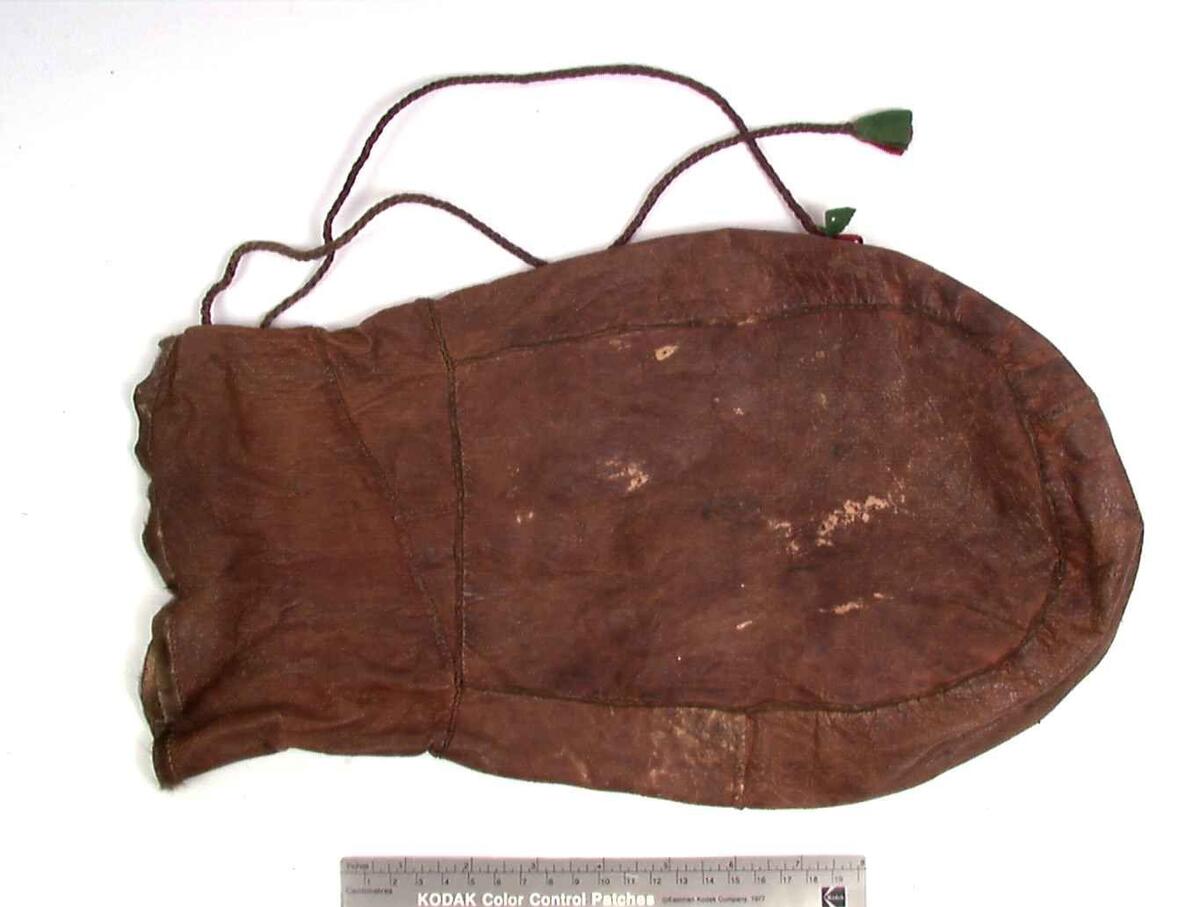 Rødbrun, avlang skinnpose med avrundet bunn, med glatthåret pelskant rundt åpningen og 2 tvunnede snorer med dusker av røde og grønne tekstilstykker.