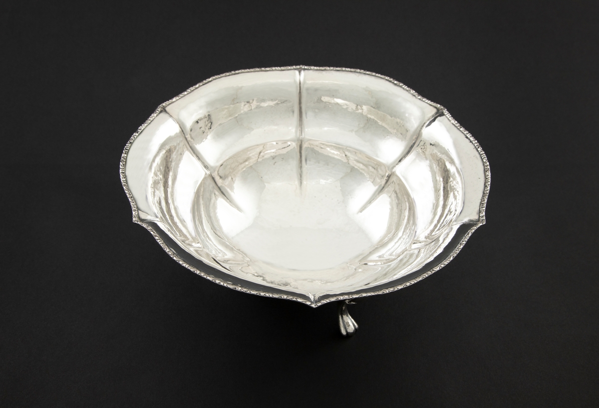 Sølvskål med rund, dyp form, bølgende kant og rette knekk. Den står på tre svungne bein. Kanten er dekorert med bonderomantisk ornamentikk.