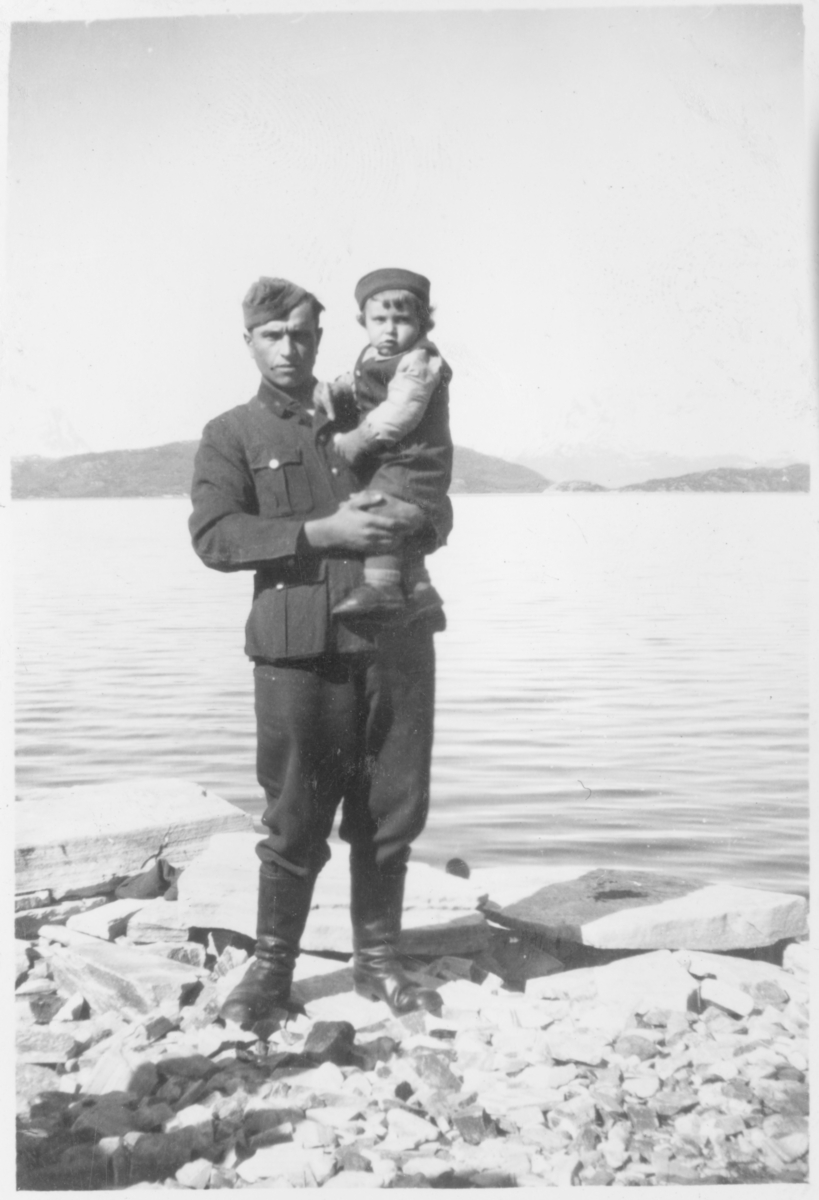 Mann i uniform med en liten gutt på armen. Fjord i bakgrunnen.