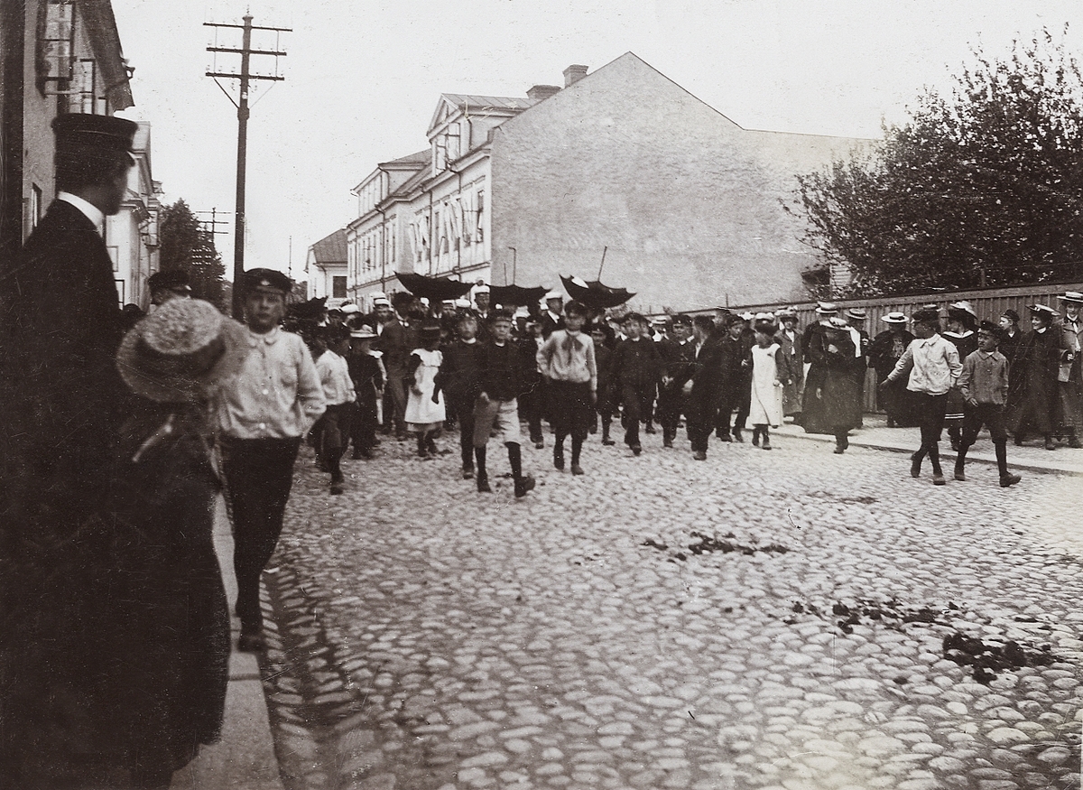 Studentupptåg i Växjö, ca 1910. De nybakade studenterna är på väg längs Västergatan mot mössmakare Lindahl på Västergatan 1, med upp-och-ner vända paraplyer.