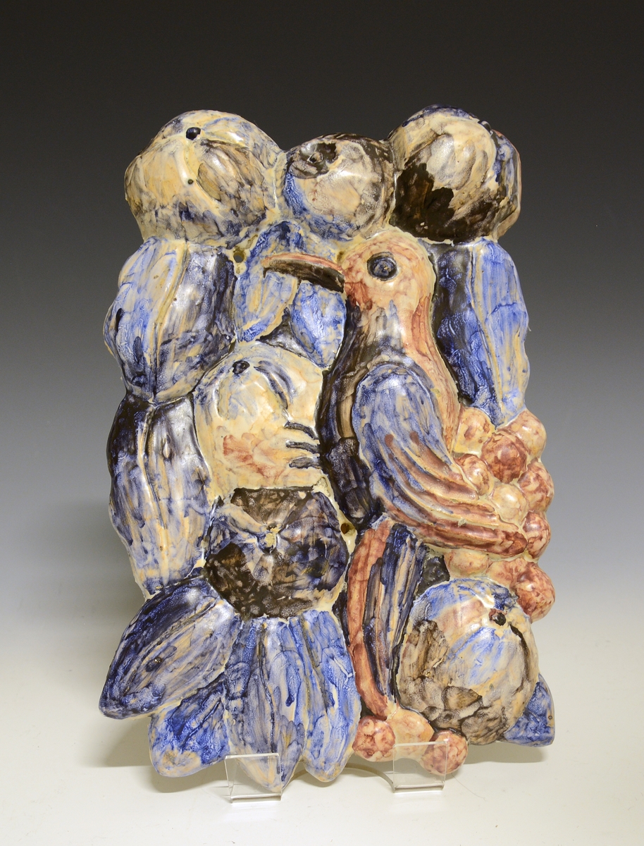 Prot: Relieff av "keramikk". Motiv: Fugl og frukt. Grovt modellert og fargelagt i blå og brune farger. To hull i godset for skruefeste. Merket "ppk nora g. 1944"