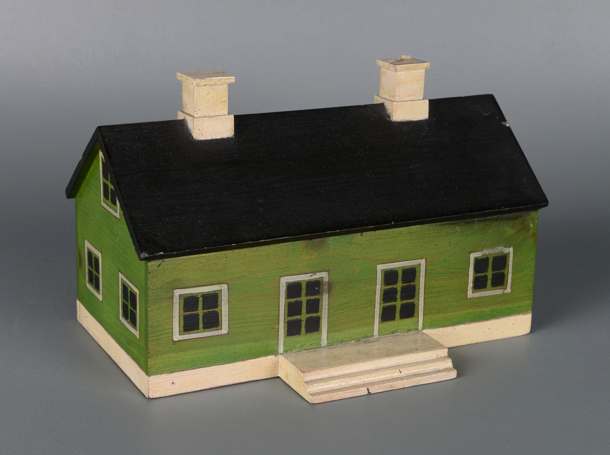 En liten modell av et hus laget av re og montert på en treplate. Huset er rektangulært med skrått tak. Det har to piper, tre dører og 12 vinduer og en liten trapp. Huset er malt grønt, med hvite karmer og hvite piper.