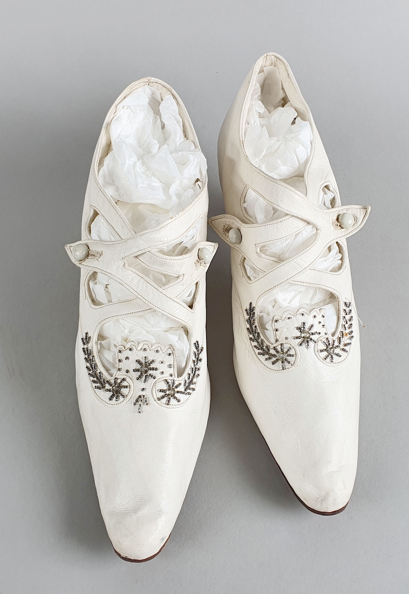 Hvite brudesko av mykt skinn med reimer og dekor av sølvperler over vristen, og med knapp på hver side. Skoene har middels høye hæler som skråner inn under sålen.
